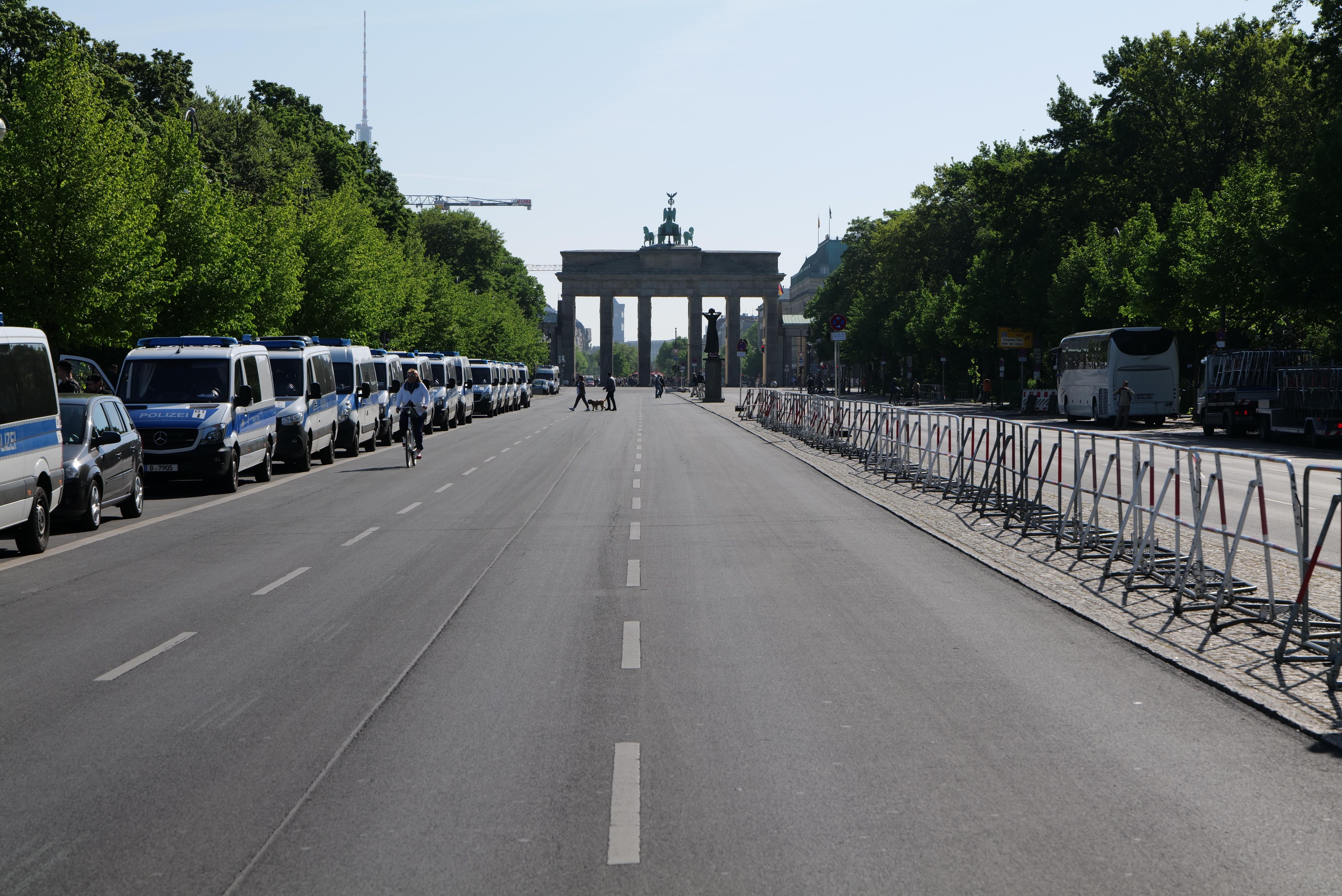 Lange Reihe von Polizeiwagen auf der Straßenseite vor dem Brandenburger Tor in Berlin, Deutschland geparkt, mit Menschen auf Fahrrädern und auf der Straße stehend, Barrieren, Bäume, ein Bogen mit Statuen im Hintergrund und sichtbarem Himmel.