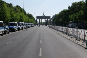 Lange Reihe von Polizeiwagen auf der Straßenseite vor dem Brandenburger Tor in Berlin, Deutschland geparkt, mit Menschen auf Fahrrädern und auf der Straße stehend, Barrieren, Bäume, ein Bogen mit Statuen im Hintergrund und sichtbarem Himmel.