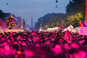 Eine große Menge bei einem nächtlichen Musikfestival mit bunten Lichtern und Ballons, umgeben von Zelten, Bannern, Fahnen, Bäumen, einem Turm und einem sternenklaren Himmel.