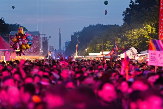 Eine große Menge bei einem nächtlichen Musikfestival mit bunten Lichtern und Ballons, umgeben von Zelten, Bannern, Fahnen, Bäumen, einem Turm und einem sternenklaren Himmel.