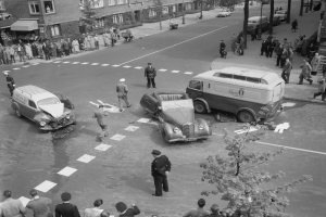 Schwarz-weiß-Szene eines Autounfalls an einer Straßenkreuzung mit mehreren Fahrzeugen, darunter ein Lieferwagen, und einer Gruppe von Schaulustigen; Hintergrund zeigt Gebäude, Bäume, Strommasten und Texttafeln.