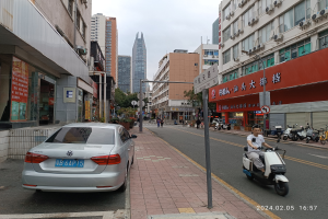 Ein Mann fährt mit einem Roller eine Straße in der Stadt entlang, mit Fußgängern auf den Bürgersteigen und anderen Fahrzeugen auf der Straße, unter einem klaren blauen Himmel mit Bäumen im Hintergrund.