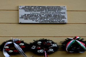 Zwei Kränze mit Bändern hängen an der Seite eines Gebäudes, mit einer Holocaust-Gedenktafel an der Wand dahinter.