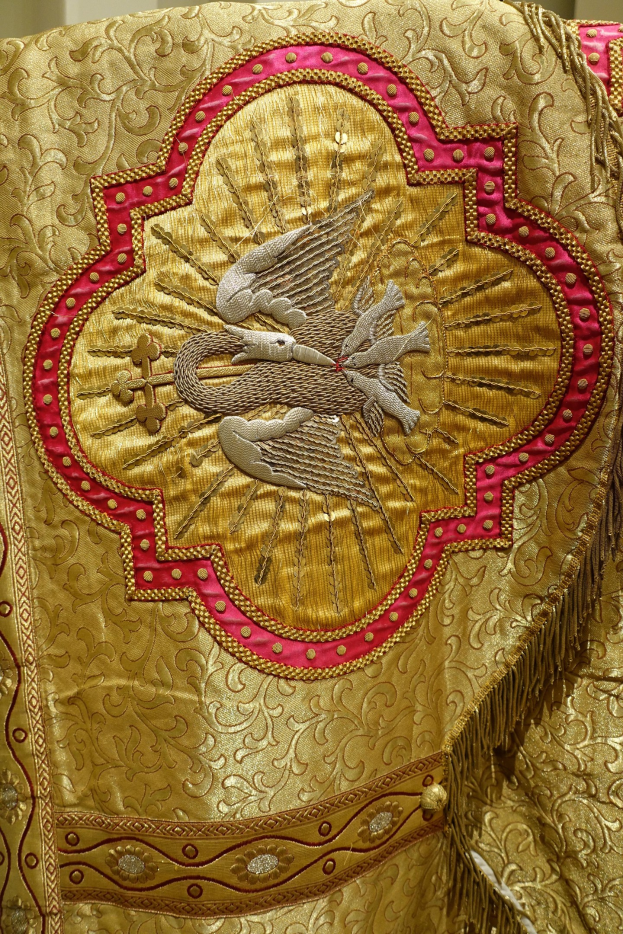 Nahaufnahme eines Priestergewandes mit goldenen und roten Stickereien und detaillierten Mustern, vor einer Wand im Hintergrund.