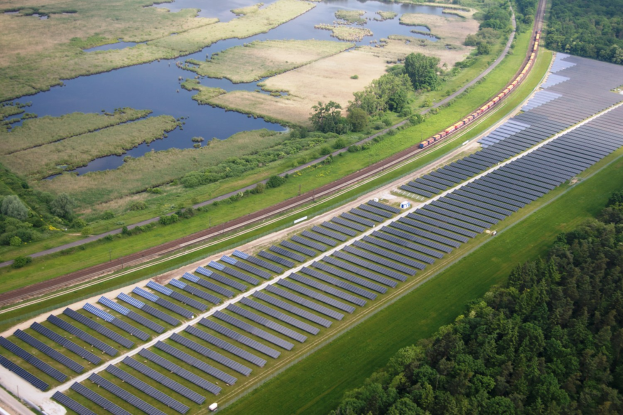 Luftaufnahme einer Solar-Farm mit Solar-Panelen in einem Feld, umgeben von Bäumen, Gras, Pflanzen und Wasser, mit einem Zug auf einer nahen Bahnschiene.