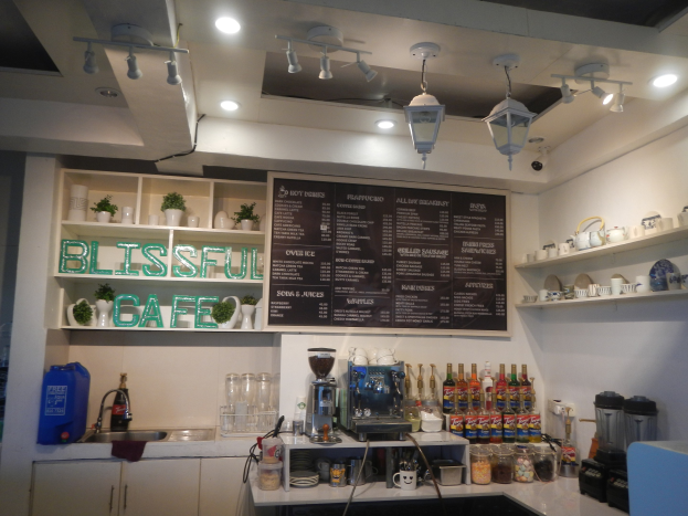 Ein gemütliches Cafe-Interieur mit einer Theke, die Flaschen, Tassen und Küchenutensilien zeigt, eine Speisekarte an der Wand, Schränke und Regale mit Tellern und Geschirr im Hintergrund, ein Waschbecken links und eine warme Atmosphäre durch Deckenleuchten.