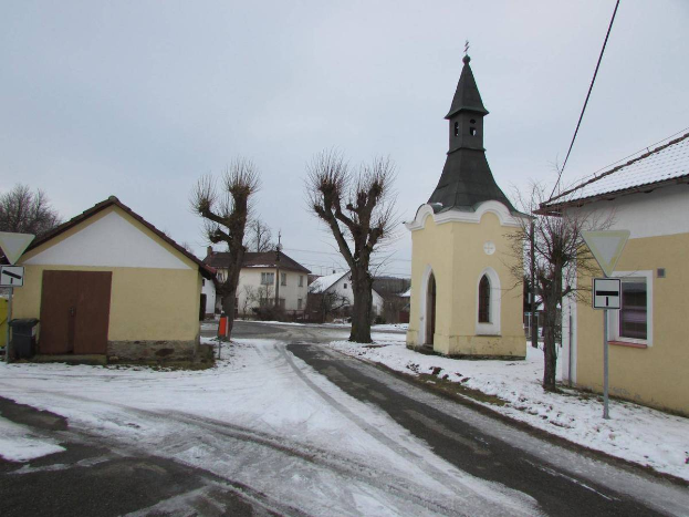 Ein kleines Dorf mit einer Kirche in der Mitte, umgeben von Häusern, Schildern, einem Müllcontainer, Bäumen und Drähten, alles von Schnee bedeckt unter einem bewölkten Himmel.
