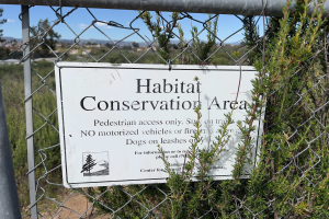 Ein Schild an einem Maschendrahtzaun mit der Aufschrift "Habitat-Schutzgebiet" umgeben von Pflanzen, Bäumen, Hügeln und einem klaren blauen Himmel.