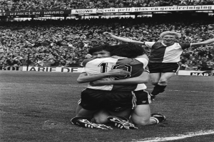 Drei Fußballspieler feiern ein Tor auf dem Boden, indem sie sich gegenseitig umarmen, vor einem jubelnden Publikum und schwenkenden Fahnen im Hintergrund, dargestellt in Schwarz-Weiß.