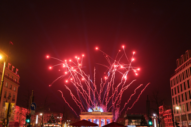 Eine belebte Stadtstraße bei Nacht am Silvesterabend in Berlin, mit Gebäuden, Bäumen, Laternenmasten, Verkehrsampeln, Schildern, Zelten, Menschen und einem prächtigen Feuerwerk am Himmel.
