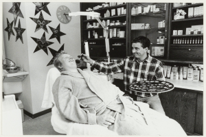 Ein Mann sitzt lachend in einem Zahnarztstuhl, während ein anderer Mann neben ihm steht und ein Tablett hält, mit einem Waschbecken, Sternen, einer Uhr und Regalen voller Flaschen im Hintergrund.