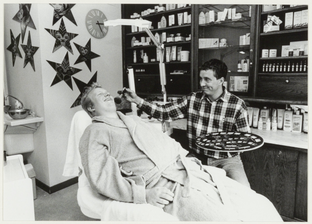Ein Mann sitzt lachend in einem Zahnarztstuhl, während ein anderer Mann neben ihm steht und ein Tablett hält, mit einem Waschbecken, Sternen, einer Uhr und Regalen voller Flaschen im Hintergrund.