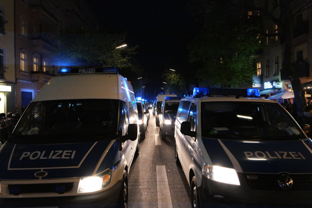 Eine Gruppe von Polizeiwagen auf einer Straße bei Nacht geparkt, mit Gebäuden, Bäumen und dunklem Himmel im Hintergrund und Menschen und Zelten auf der rechten Seite.