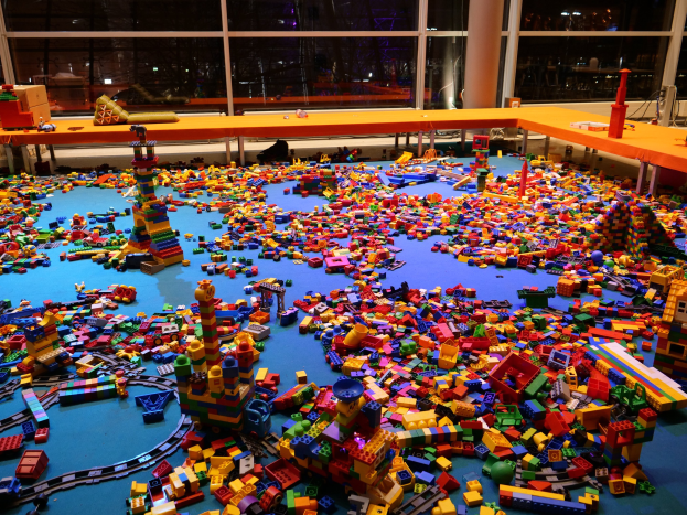 Ein großer Raum mit farbigen Lego-Steinen, die auf dem Boden verstreut sind, umgeben von Tischen und Säulen, mit Glasfenstern im Hintergrund, die Menschen und Lichter zeigen.