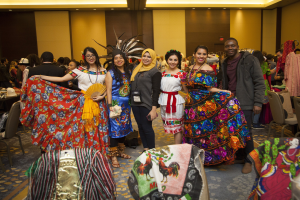 Eine Gruppe von Menschen in bunten Kleidern, die fr├Âhlich posieren und fotografiert werden, bei einer mexikanischen Kulturveranstaltung mit Stühlen, Tischen und Kleidung im Hintergrund.