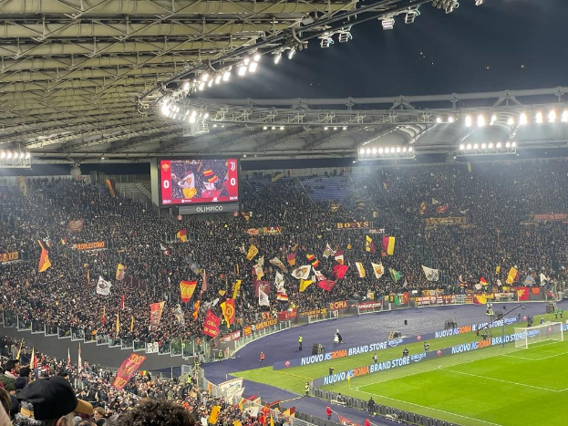 Eine große Menge Menschen in einem hell erleuchteten Stadion schwenkt Fahnen und Banner und wartet gespannt auf ein Fußballspiel, wobei Rasen, Netz und eine Leinwand im Hintergrund zu sehen sind.