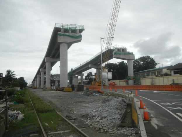Baustelle mit Brücke, Straße mit Absperrbaken, Bahnschiene, Steine, Gras, Bäume, Gebäude und bewölkter Himmel.