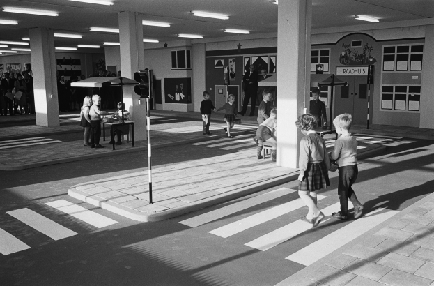 Schwarzes und weißes Foto von Kindern, die eine Straße an einer Ampel überqueren, mit Fußgängern in der Nähe, einem Verkehrssignalmast im Vordergrund und Hintergrundsäulen mit Deckenleuchten.