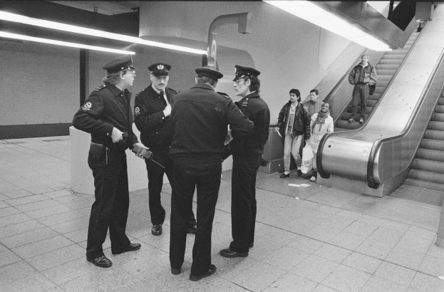 Schwarzes und weißes Bild von mehreren Polizeibeamten, die zusammen in einer U-Bahn-Station stehen, mit einer Rolltreppe auf der rechten Seite und Deckenlampen darüber.