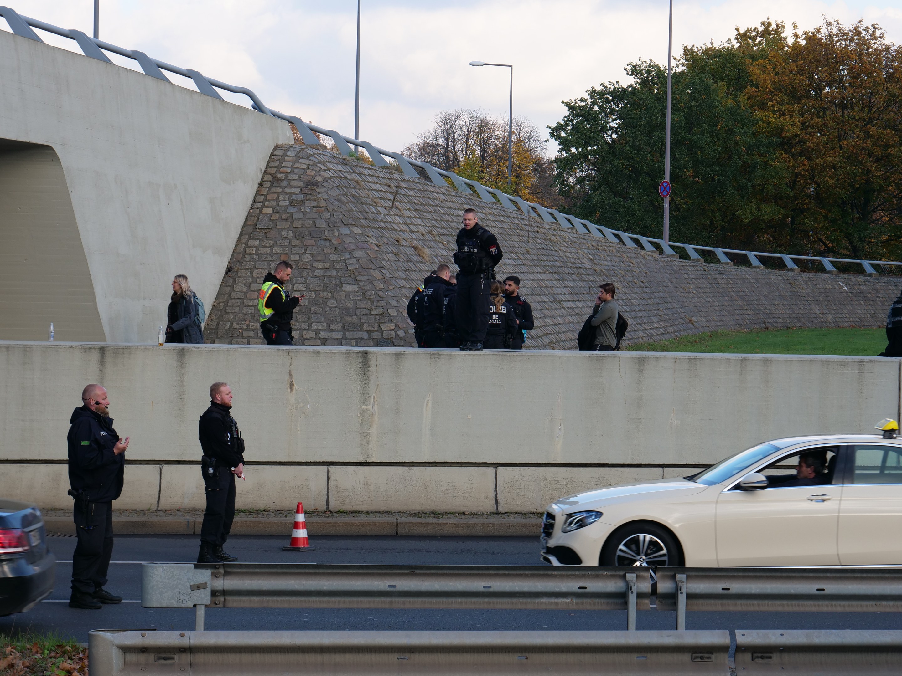 Gruppe von Polizisten, die neben einem Auto auf der Seite einer Straße stehen, mit Verkehrskegeln, einer Trennwand, Gras, einer Wand, Laternenmötzen, Bäumen und einem bewölktem Himmel im Hintergrund.