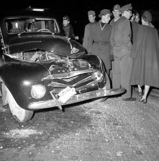 Schwarz-weiß-Bild eines schwer beschädigten Autos am Straßenrand mit eingedellter Motorhaube und zersplitterten Fenstern, umgeben von Menschen, die die Szene begutachten.