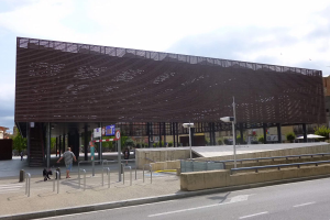 Großes modernes Gebäude mit zahlreichen Fenstern, das als neuer Eingang zum Stadtzentrum dient, umgeben von Straßeninfrastruktur, Fußgängern, Fahrzeugen, Bäumen und einem bewölkten Himmel.