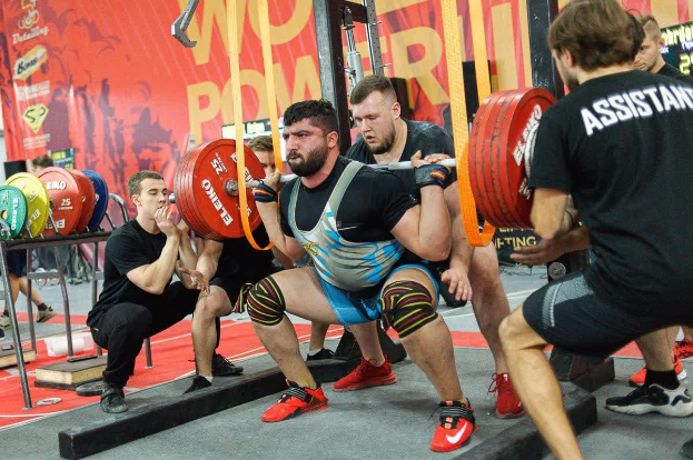 Eine Gruppe von Männern führt Kniebeugen mit Hanteln in einem mit Geräten und Bannern vollgestellten Fitnessstudio durch, auf denen "Powerlifting-Weltmeisterschaften 2019" steht.