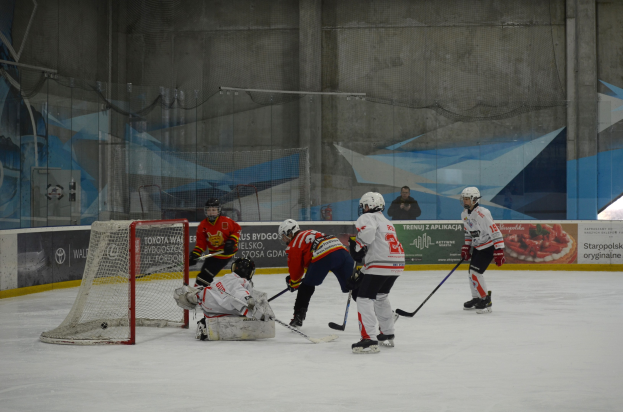 Gruppe von Menschen, die Eis-hockey auf einem Eisplatz spielen, mit Helmen und Hockey-Schlägern, mit einem Torpfosten auf der linken Seite, Bannern im Hintergrund und einer Wand.
