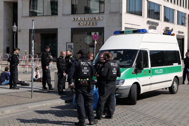 Eine Gruppe von Polizeibeamten steht vor einem Starbucks-Café, rechts davon parkt ein Van und links stehen und sitzen einige Menschen, im Hintergrund sind ein Gebäude, ein Schild, ein Laternenmast und ein Zaun zu sehen.