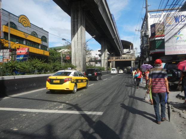 Gelbes Taxi fährt auf einer Straße mit hohen Gebäuden, Fußgängern auf dem Gehweg mit Regenschirmen, einer Brücke im Hintergrund, bewölktem Himmel und Strommasten mit Überlandleitungen.