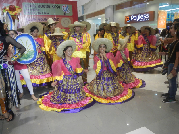 Eine Gruppe von Menschen in bunten Kleidern und Hüten tanzt in einem Einkaufszentrum, während andere Musikinstrumente halten und ein Banner tragen, bei einer fröhlichen Cinco de Mayo-Feier.