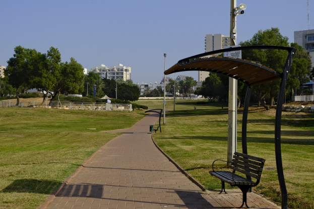 Eine Parkbank auf einem grünen Feld mit Bäumen, Masten, Lichtern und Gebäuden im Hintergrund und einem Mülleimer in der Vordergrund.