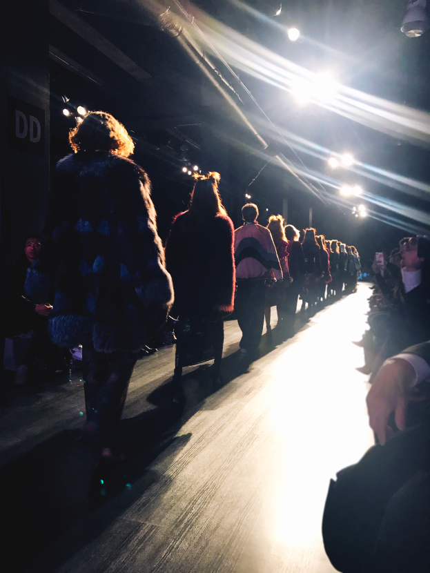 Eine Gruppe von Menschen läuft über einen hell erleuchteten Laufsteg bei einer Modenschau, während Zuschauer auf beiden Seiten sitzen.