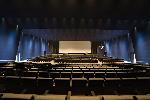 Ein großer Zuschauerraum mit Reihen von Stühlen, wandmontierten Lampen, einer Projektionsleinwand und ein paar Gegenständen auf der Bühne.