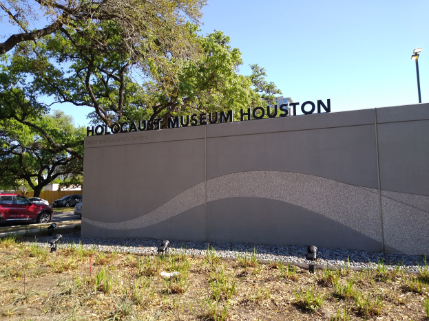 Außenansicht des Holocaust-Museums Houston mit eingravierter Textwand, verstreuten Steinen, Grünfläche, Bäumen, vorbeifahrenden Fahrzeugen, einem Straßenpfahl und einem bewölkten Himmel.