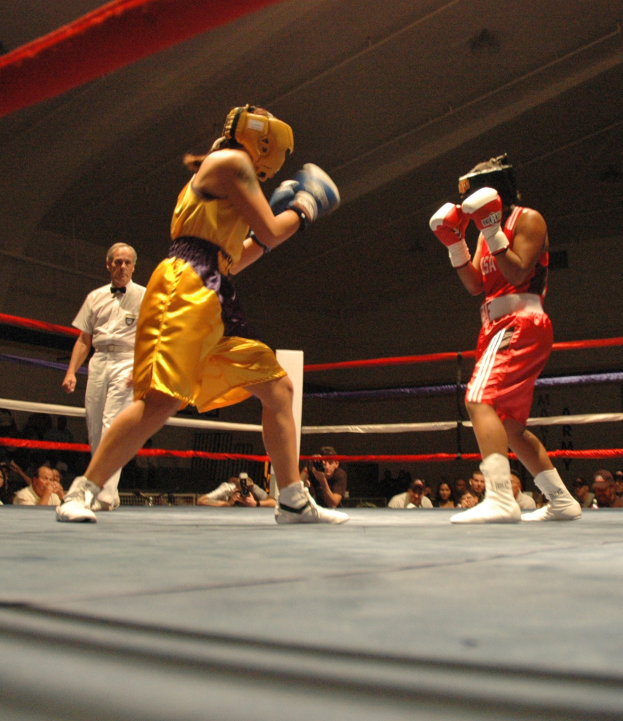 Zwei Boxer, die in einem Ring mit einem Schiedsrichter und Zuschauern, einige mit Kameras, unter einer sichtbaren Decke kämpfen.
