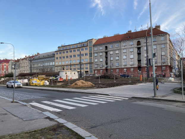 Baustelle für ein neues Wohnhaus im Vordergrund einer Stadtstraße mit parkenden Autos, Laternen, Bäumen, Gebäuden mit Fenstern und einem bewölkten Himmel.