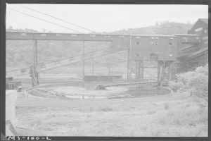 Ein Schwarz-Weiß-Foto eines Kohlebergwerks mit einer Brücke, Pfählen, Drähten, Gebäuden, Bäumen, Pflanzen, Gras und Wasser im Vordergrund und einem Himmel im Hintergrund sowie einem Text unten.