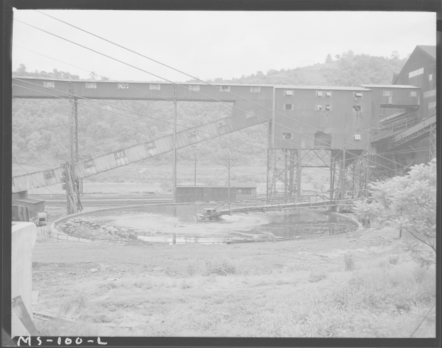 Ein Schwarz-Weiß-Foto eines Kohlebergwerks mit einer Brücke, Pfählen, Drähten, Gebäuden, Bäumen, Pflanzen, Gras und Wasser im Vordergrund und einem Himmel im Hintergrund sowie einem Text unten.