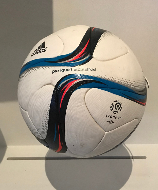 Ein weißer Adidas-Fußball mit schwarzer 'Ligue 1'-Aufschrift liegt auf einem weißen Tisch vor einem hellen weißen Hintergrund.