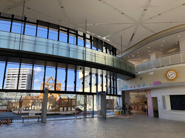 Innenansicht eines großen Gebäudes mit zahlreichen Fenstern, Deckenbeleuchtung, einer Wanduhr, einem Fernseher, einem Anzeigebildschirm, Holzkonstruktionen einschließlich eines Zauns, entfernte Gebäude, Bäume und einen bewölkten Himmel, der den Eingang zu einem neuen Kinderspielplatz markiert.