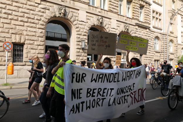 Eine Gruppe von Menschen marschiert auf einer Stadtstraße, einige halten Schilder und andere fahren Fahrräder, im Hintergrund ein historisches Gebäude mit rundbogigen Fenstern, Säulen und Skulpturen.
