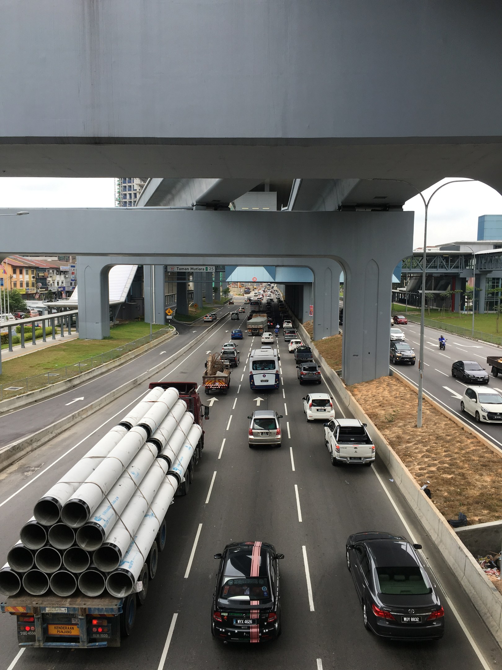 Schwerer Verkehr auf einer Autobahn unter einer Brücke mit Straßenlaternen, Gras, Gebäuden, Bäumen und dem Himmel im Hintergrund.