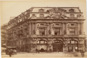 Schwarz-weiß-Foto des Théâtre de la Renaissance in Paris mit einer Fassade mit Fenstern, Säulen, Bögen und Skulpturen, mit ein paar Menschen und Fahrzeugen auf der Straße davor und dem Himmel im Hintergrund.