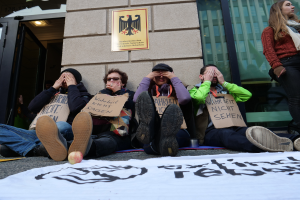 Eine Gruppe von betroffenen Menschen sitzt auf dem Boden und hält Schilder, mit einer Fahne, die Text und einen Apfel trägt, auf der Straße, und eine Frau steht rechts daneben, vor einem Gebäude mit Glasfenstern und einer Tür, und einer Wand mit einer Tafel im Hintergrund.