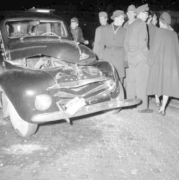 Schwarz-weißes Bild eines schwer beschädigten Autos am Straßenrand mit eingedellter Motorhaube und zersplitterten Fenstern, umgeben von einer Gruppe von Menschen, die es besorgt betrachten.