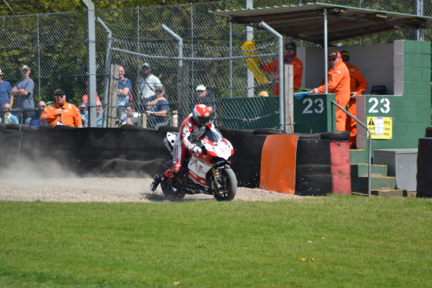 Ein Motorradfahrer in Helm und Handschuhen fährt über ein grasbewachsenes Feld, mit Zuschauern in Kappen und Brillen, einem Zaun, Treppen mit Geländern und einem nummerierten Schuppen im Hintergrund.