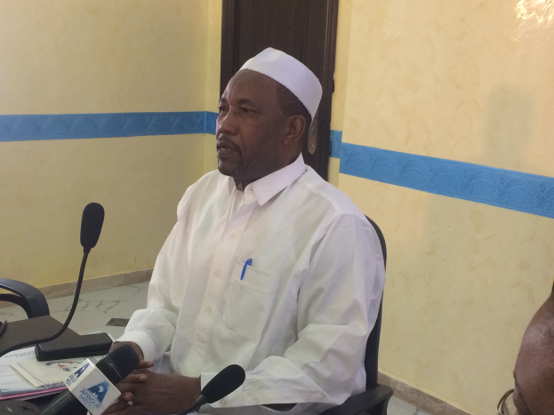 Ein Mann in einem weißen Gewand und Hut sitzt an einem Tisch mit Mikrofonen, spricht in einem Raum mit einer anderen Person zu seiner Rechten, Papiere auf dem Tisch und eine Tür im Hintergrund.