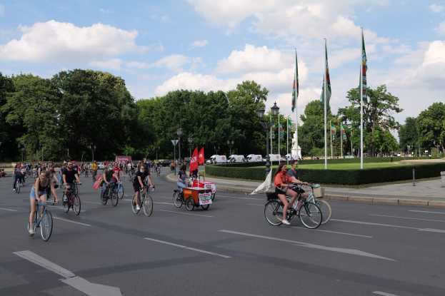 Eine Gruppe von Menschen, die auf Fahrrädern eine von Bäumen gesäumte Straße mit Laternenmasten und Fahnenmasten entlangfahren, während der Pariser Radtour 2016, mit Fahrzeugen und bewölktem Himmel im Hintergrund.