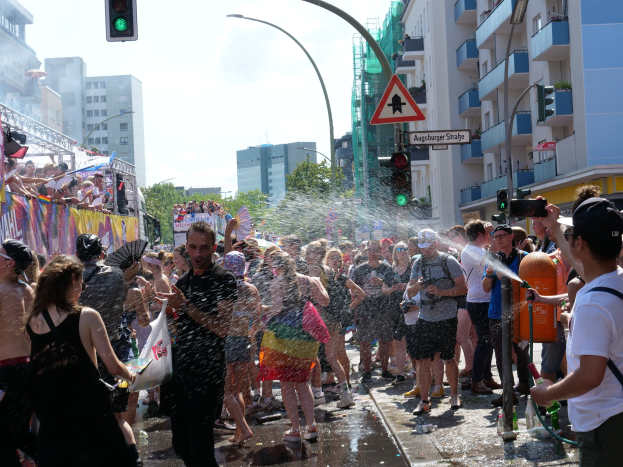 Menschen auf einer Christopher Street Day Parade, die sich gegenseitig mit Wasser bespritzen und Gegenstände halten, mit einem Banner links und Gebäuden, Bäumen und Ampeln im Hintergrund.